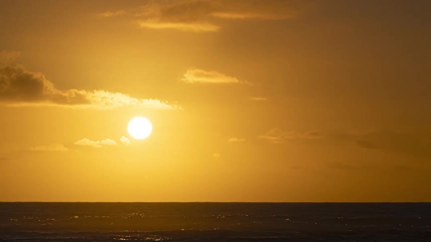 A serene time-lapse captures the sun rising from the ocean horizon, illuminating the sky with a warm, golden light