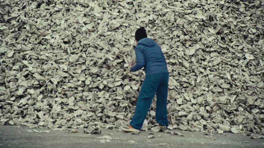 A woman in a blue jacket inspects a shell from a giant mound of discarded oyster shells and walks away in slow motion