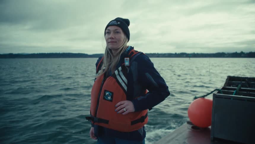 A resolute woman wearing a life vest and beanie stands on a moving boat, looking out over the water on an overcast day