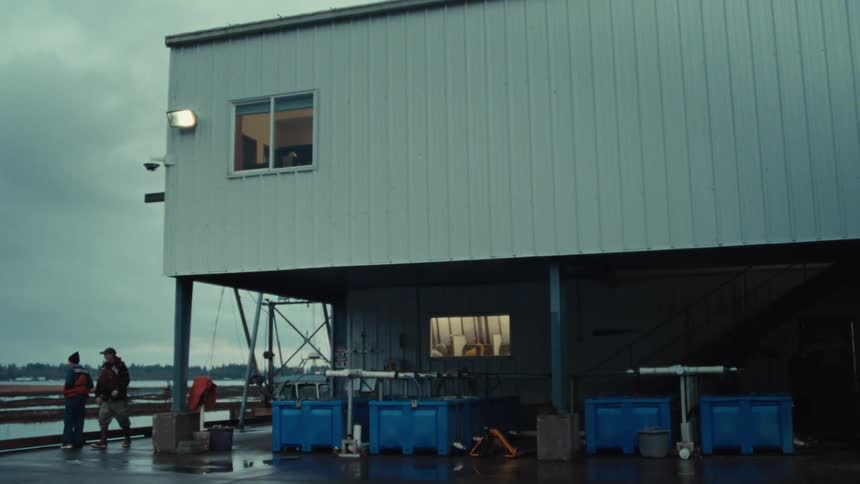 Two workers chat on a wet industrial pier next to a processing building on a gloomy day