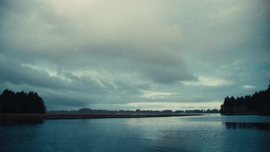 A wide, tranquil river flows through a dense forest under a moody, overcast sky