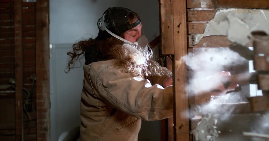 A construction worker wearing a respirator mask smashes through a plaster wall with a sledgehammer in super slow motion