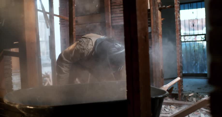 Worker wearing a respirator mask shovels rubble and dust into a wheelbarrow on a demolition site in slow motion