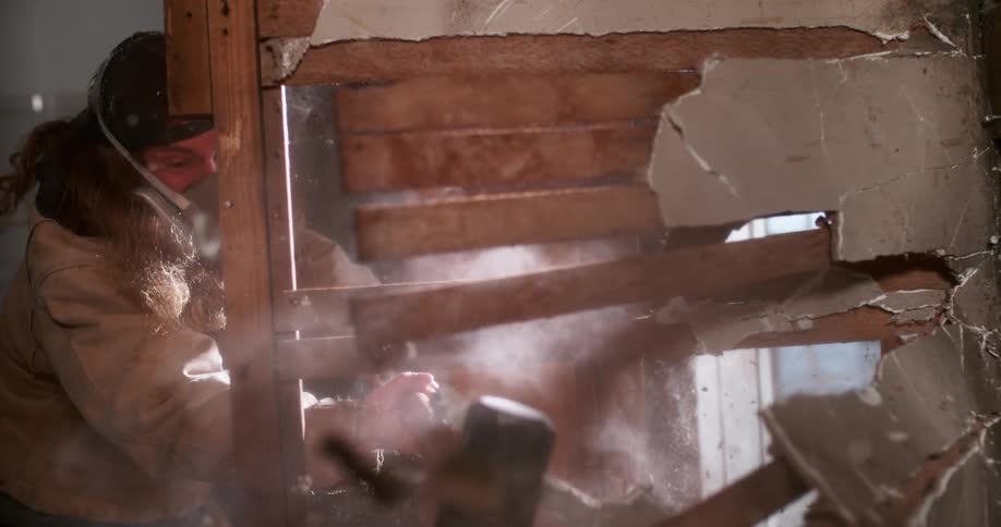 A construction worker wearing a respirator mask smashes through a plaster and lath wall with a sledgehammer in dramatic slow motion