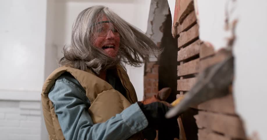 A powerful older woman in safety gear screams as she demolishes an interior wall with a sledgehammer, with plaster exploding in dramatic slow motion
