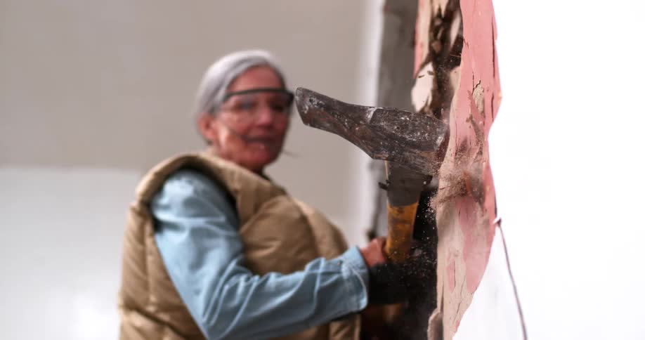 Low angle slow motion shot of a woman in safety gear swinging a large sledgehammer to demolish an interior wall