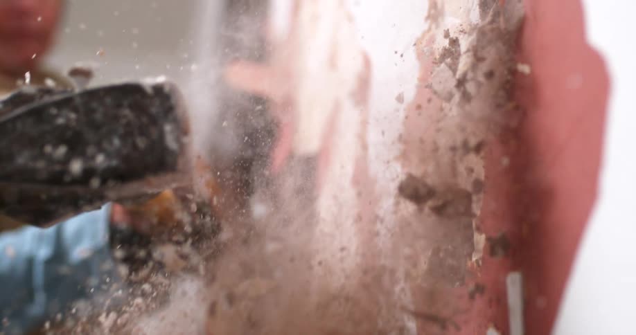 A gloved hand swings a sledgehammer, smashing a plaster wall into dust and debris in dramatic slow motion