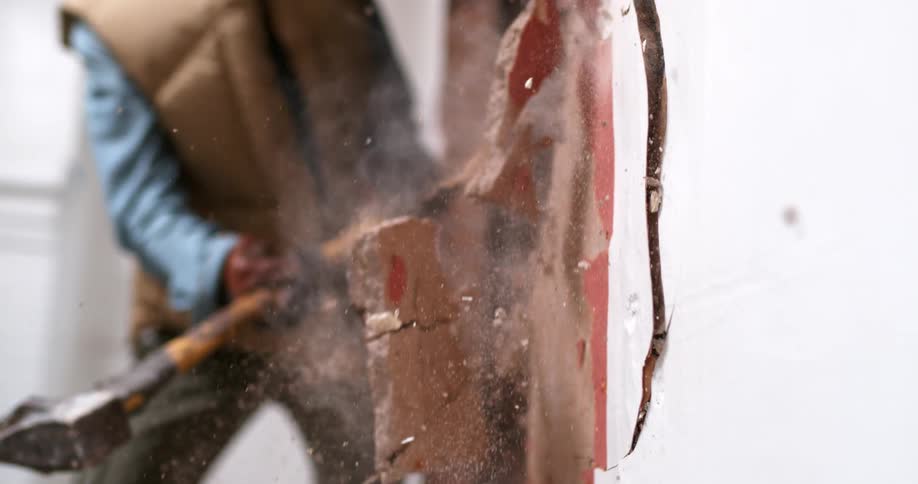 A sledgehammer smashes through a plaster wall, sending dust and debris flying in super slow motion