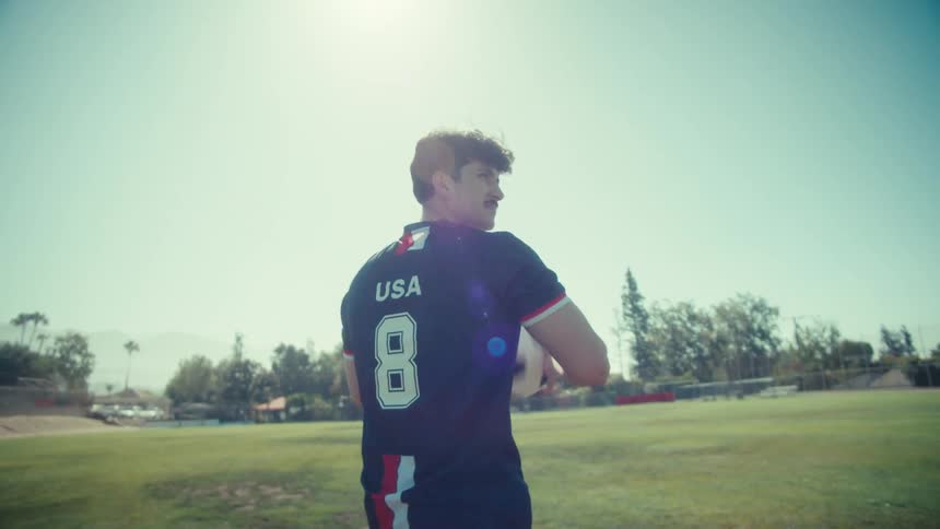 A focused young soccer player in a USA uniform holds a ball while walking across a sunny field in slow motion