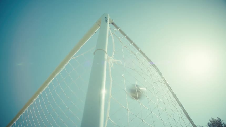 A low angle slow motion shot of a soccer ball hitting the back of the net for a goal on a sunny day