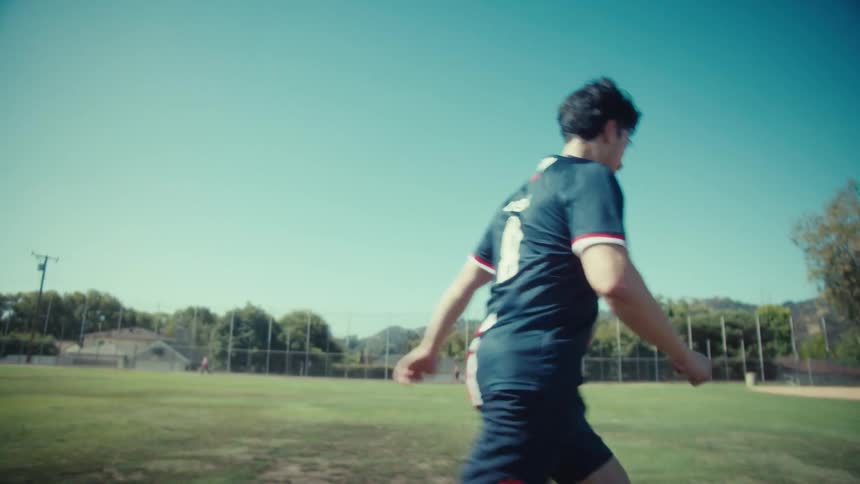 A young man in a USA soccer jersey dribbles a vintage ball down a sunny field in slow motion