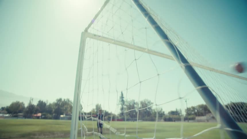 A soccer ball hits the back of the net in slow motion during practice on a sunny field as the camera pulls back