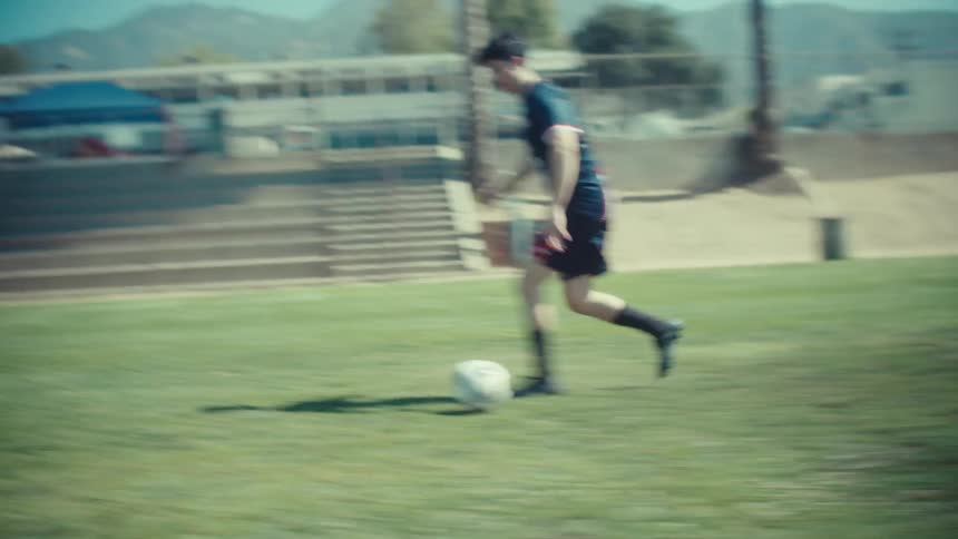 A skilled soccer player dribbles a ball down a bright green field in a dynamic slow-motion tracking shot