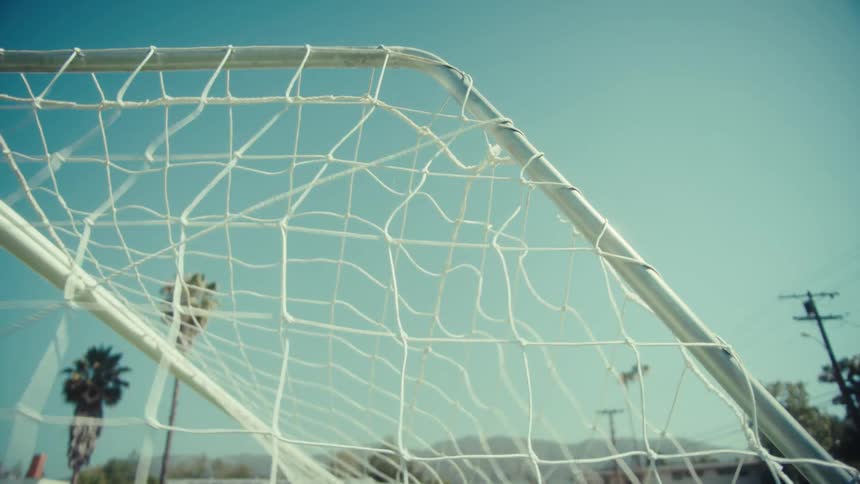 A soccer ball hits the back of the goal net in dramatic slow motion on a sunny day