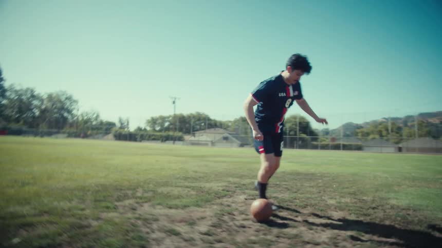 A focused soccer player in a USA jersey practices dribbling a vintage leather ball across a sunny field in slow motion before kicking it