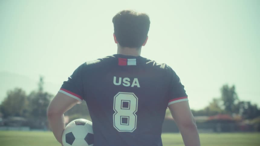 Slow motion shot from behind of a Team USA soccer player walking onto the field with a ball under his arm