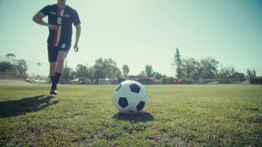 A soccer player in slow motion powerfully kicks a soccer ball on a green field on a bright sunny day