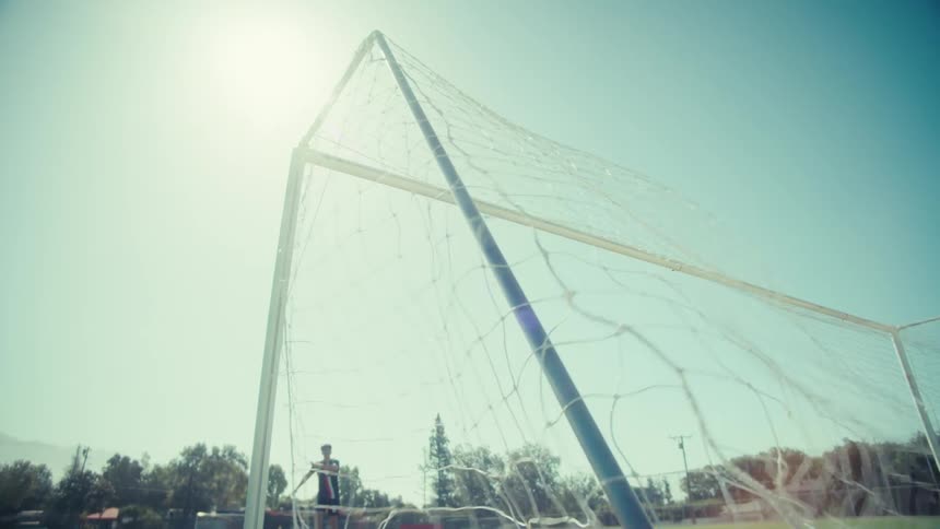 From inside the goal, a soccer ball soars into the net in slow motion on a sunny practice field
