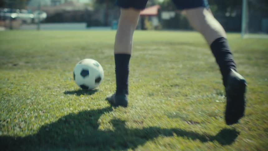 A close-up, slow-motion shot follows a soccer player's feet as they dribble a ball across a sunny grass field