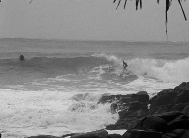 Vintage black and white footage shows surfers skillfully riding large, powerful waves on a rugged coastline