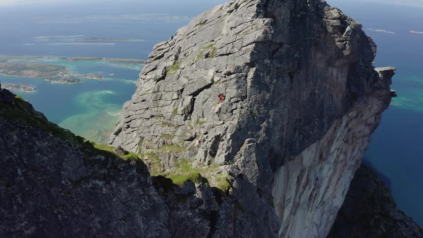 A lone rock climber scales a sheer mountain peak with an incredible aerial view of the turquoise ocean and islands below