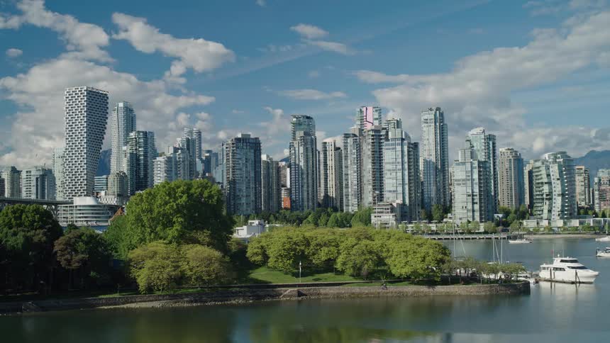 A sunny timelapse shows clouds moving across the sky over the modern Vancouver skyline, with a waterfront park and boats in the foreground