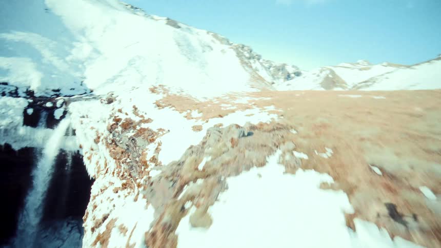 A thrilling FPV drone flight navigates a winding river through a majestic, snow-covered canyon, soaring up and over a powerful waterfall