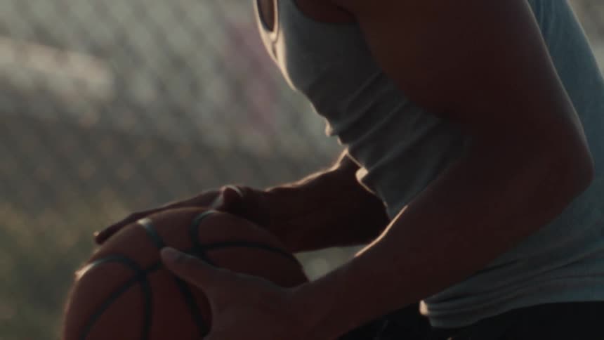 Close-up slow-motion shot of an athlete sitting and holding a basketball on an outdoor court at sunset
