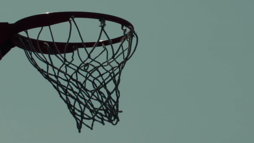 A basketball is shot through a hoop and falls through the net in slow motion against a solid color sky