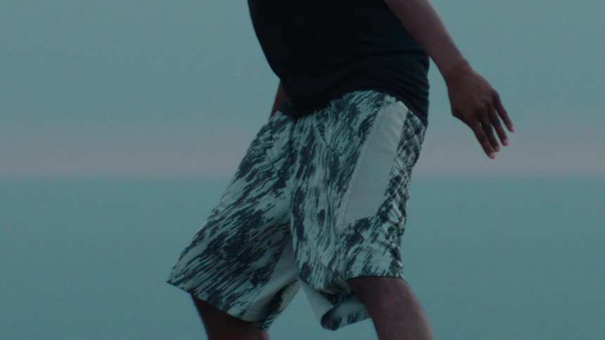 A male basketball player dribbles a ball in slow motion against the ocean horizon at dusk