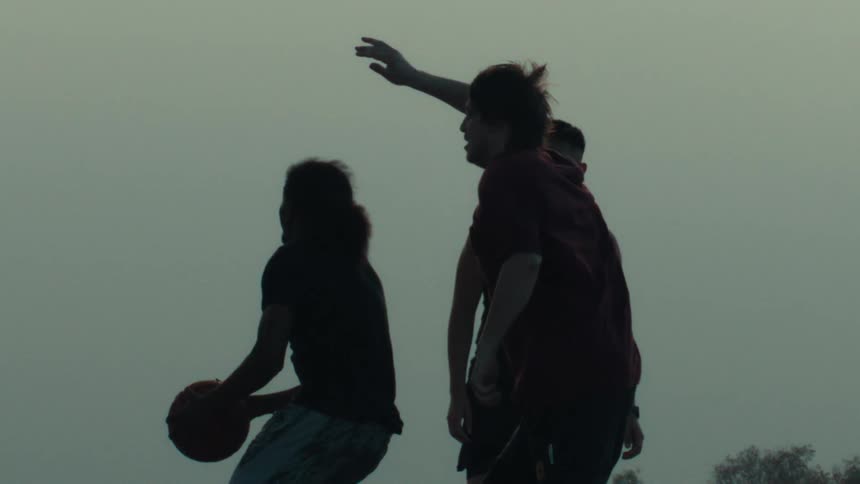 A silhouetted basketball player jumps for a layup in dramatic slow motion while being defended by his friends on an outdoor court