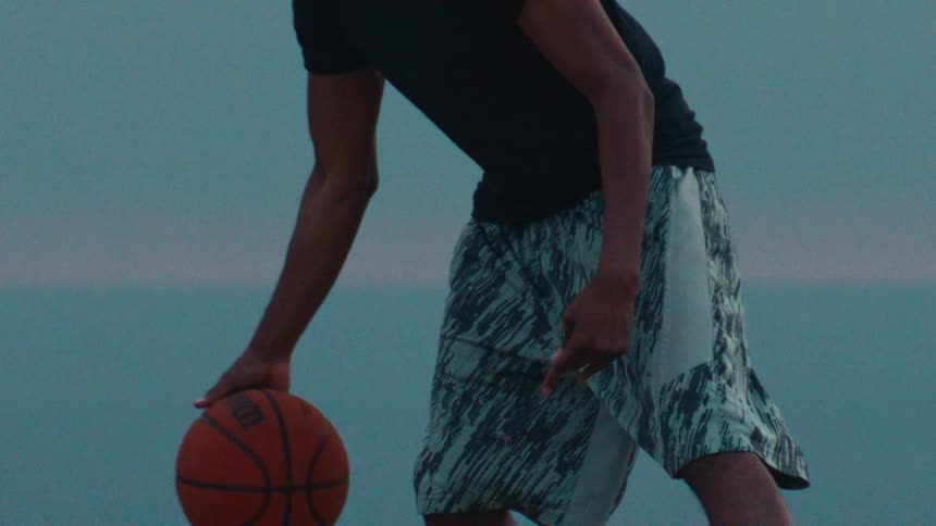 A close up in slow motion of a basketball player dribbling a ball with a calm ocean in the background at dusk