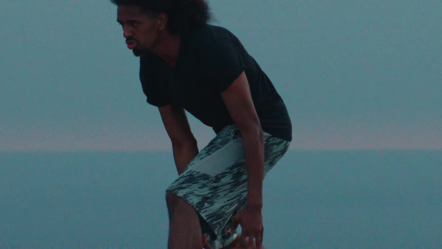 An athletic man intensely dribbles a basketball in slow motion on a grassy hill overlooking the ocean at dusk