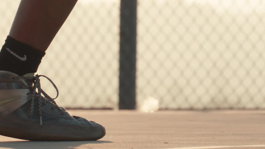 Super slow motion close up of a basketball player's shoes as they dribble the ball on a dusty court during golden hour