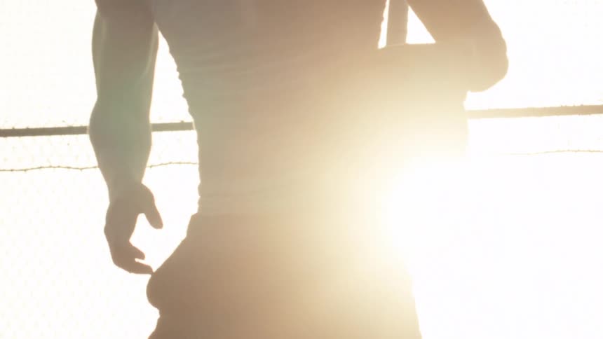 A basketball player stands silhouetted in the bright sun, holding a ball in dramatic slow motion