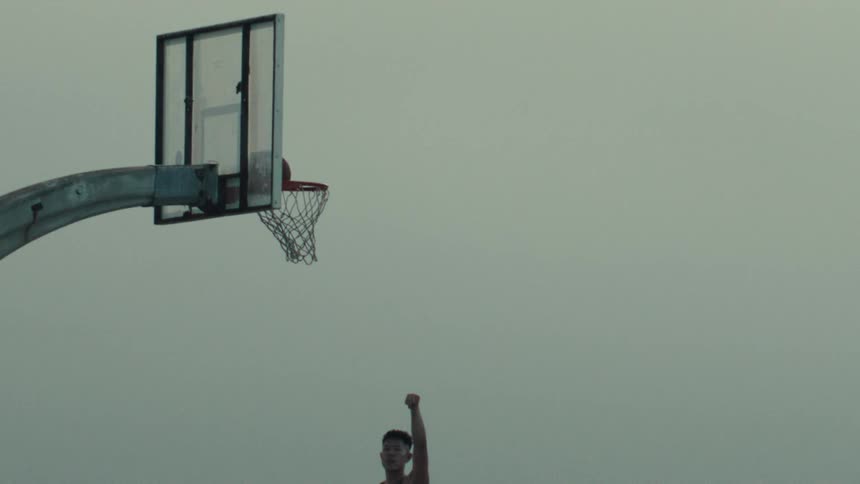 A basketball player scores a perfect basket against a grey sky in dramatic super slow motion