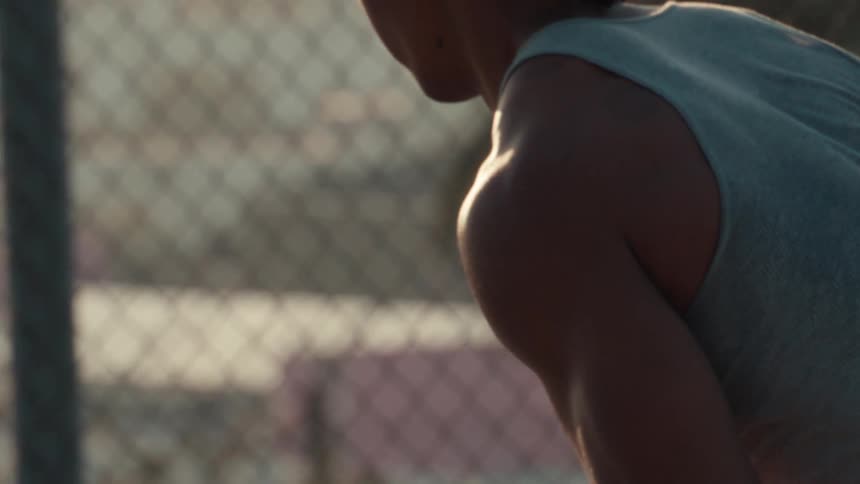 A muscular basketball player in a tank top holds a basketball in slow motion on a sunlit outdoor court with a chain-link fence