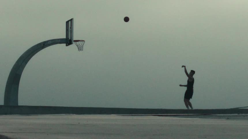 A silhouetted basketball player sinks a perfect basket in super slow motion on a hazy outdoor court