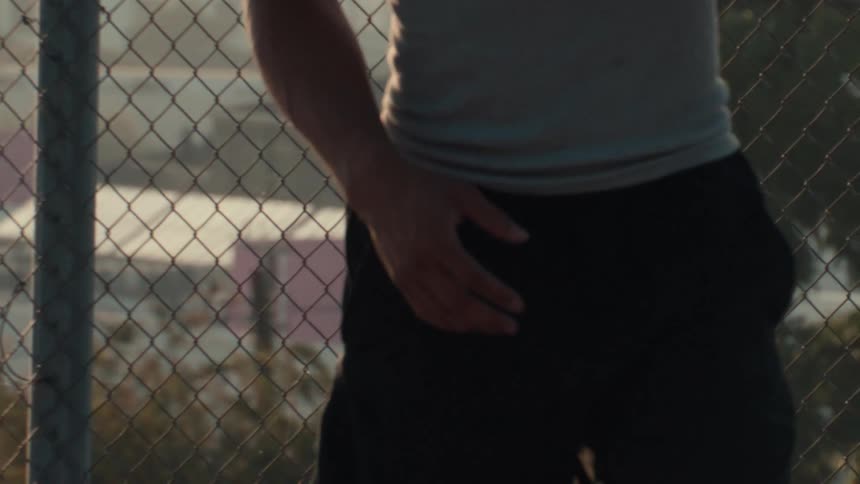 A backlit basketball player holds a ball against a chain-link fence at sunset in a slow motion close-up