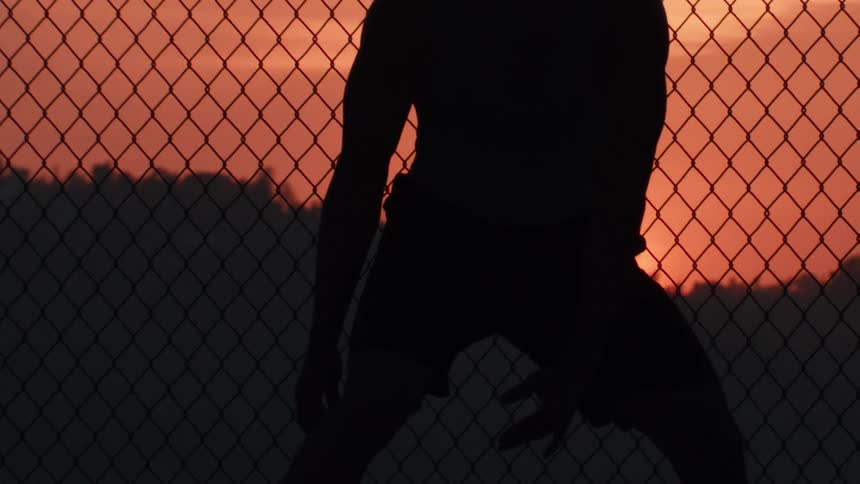 A silhouette of a basketball player dribbling in slow motion against a vibrant sunset sky behind a chain-link fence