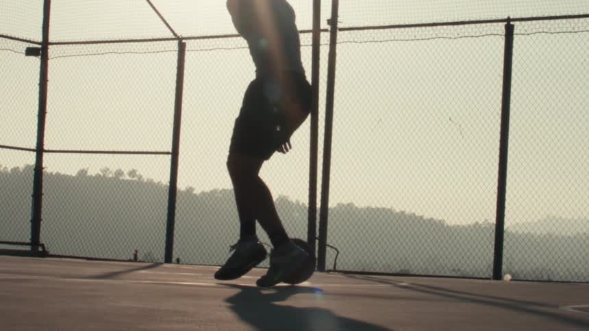 Super slow motion side view of a silhouetted basketball player dribbling on an outdoor court at sunrise