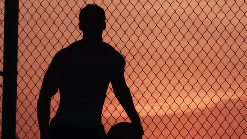 A man's silhouette dribbles a basketball in slow motion against a chain-link fence with a vibrant sunset in the background
