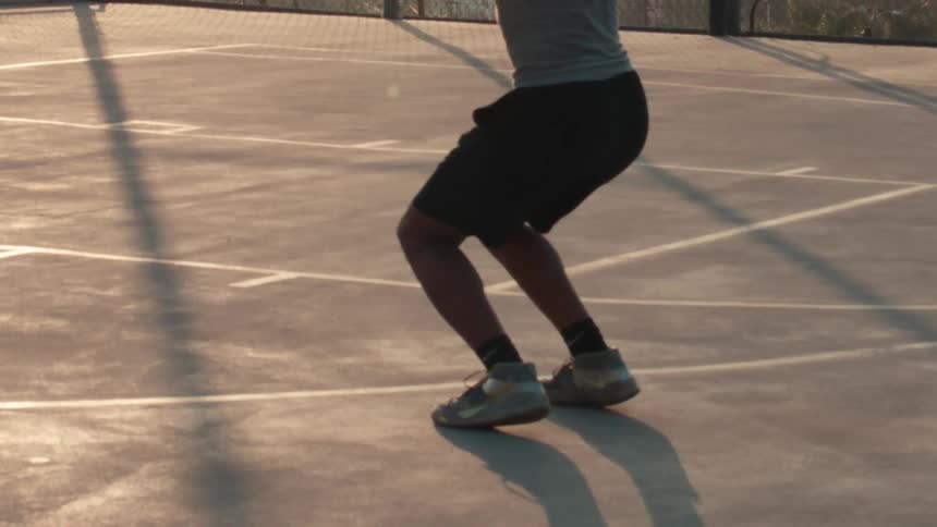 Super slow motion of a basketball player taking a jump shot on an urban court at sunrise, with the camera tilting down to focus on his athletic shoes
