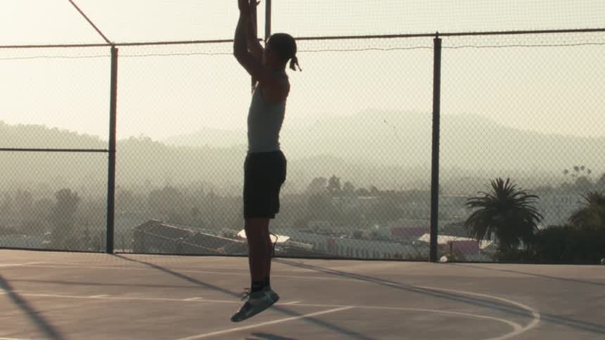 In slow motion, a silhouetted basketball player jumps to shoot a basket on an outdoor urban court at sunrise