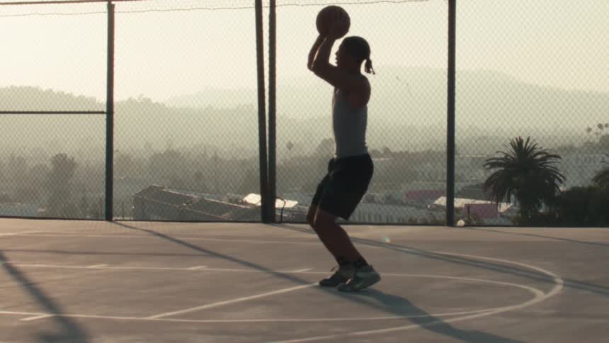 An athletic man takes a jump shot in slow motion, silhouetted against the morning sun on a city basketball court