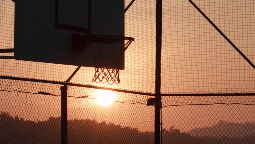 A silhouetted basketball swishes through the net in slow motion on an outdoor court during a beautiful sunset