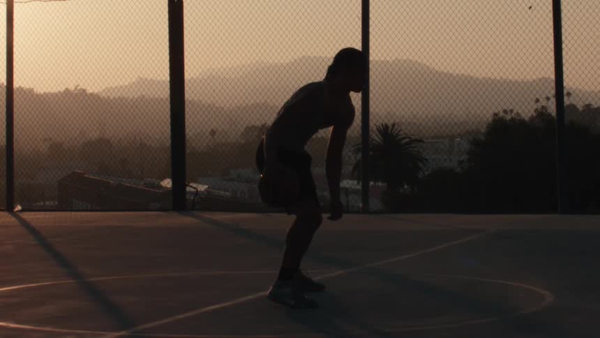 A silhouetted basketball player practices dribbling in slow motion on a scenic outdoor court during a golden sunset