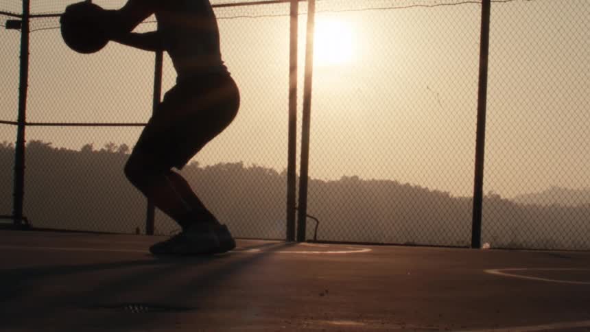 A silhouetted basketball player leaps for a jump shot in dramatic slow motion against the golden hour sun