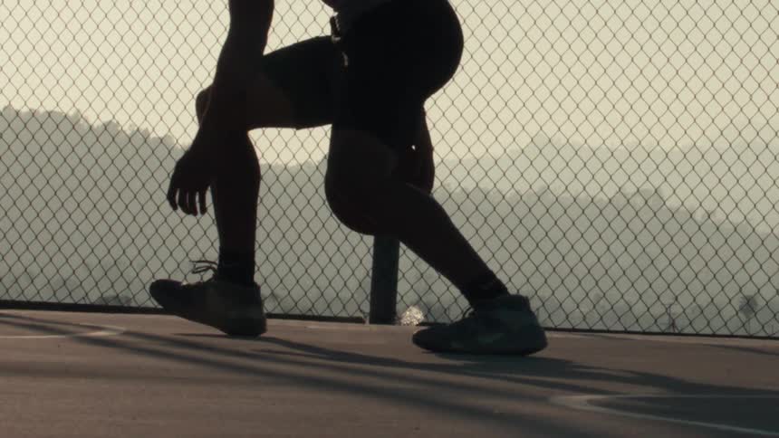 Silhouette of a basketball player practicing dribbling moves on a court with a chain-link fence in super slow motion