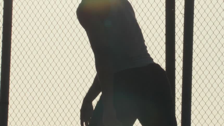 A silhouetted basketball player dribbles a ball in slow motion against a chain-link fence at sunset